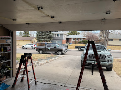 Garage interior with ladders and parked vehicles visible outside in a suburban driveway.