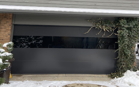 Modern garage door with frosted black glass panels framed by brick and snow-covered plants