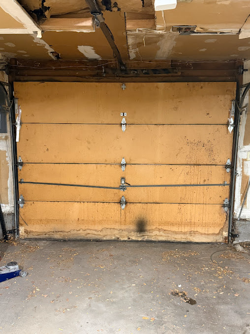 Interior of a damaged garage with a broken tan door, exposed ceiling, and debris on the floor