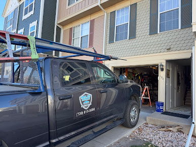 Blue pickup truck parked at an open garage of a townhouse, with a ladder rack and work tools visible.