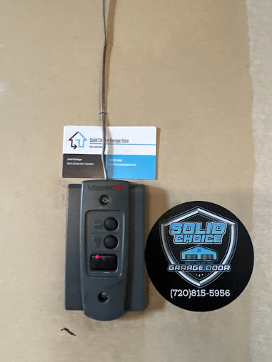 Small wall-mounted black control panel beside a business card and circular logo sticker on a beige wall.