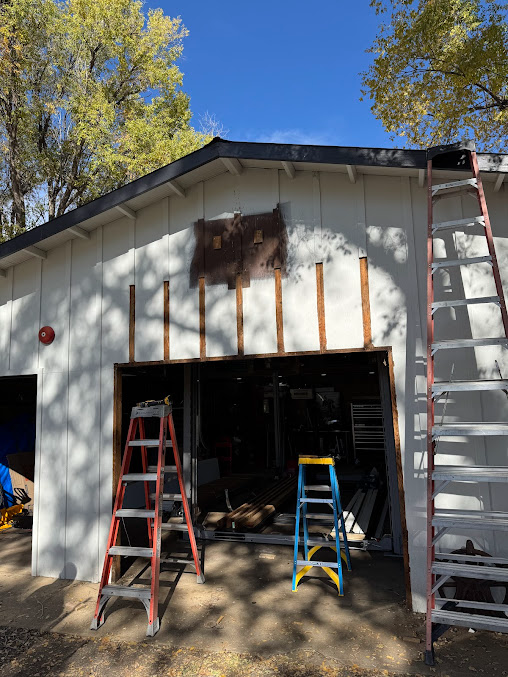 White garage under repair with ladders at the open front and on the roof, trees overhead