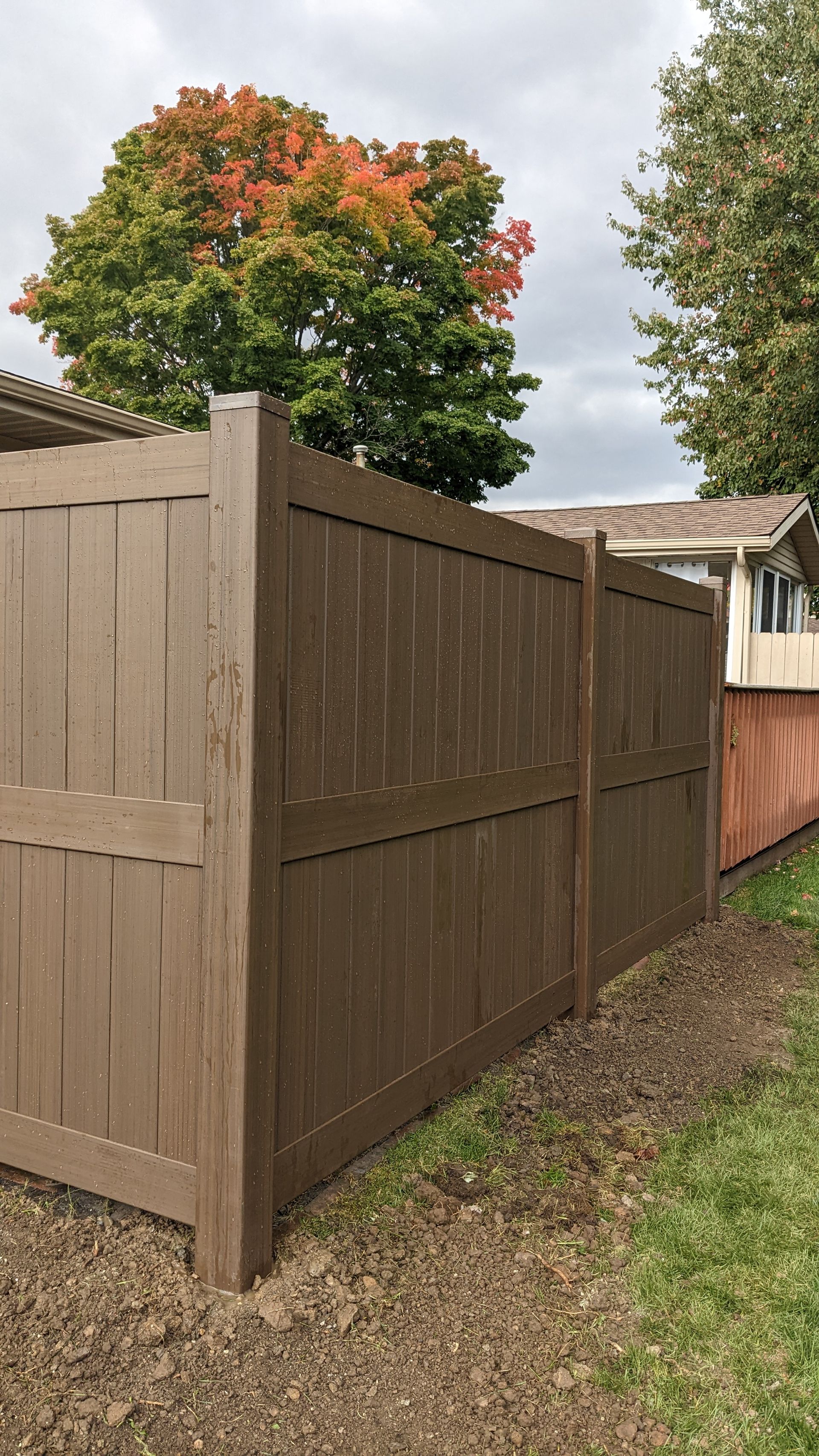 A brown wooden fence is sitting next to a house with a tree in the background.