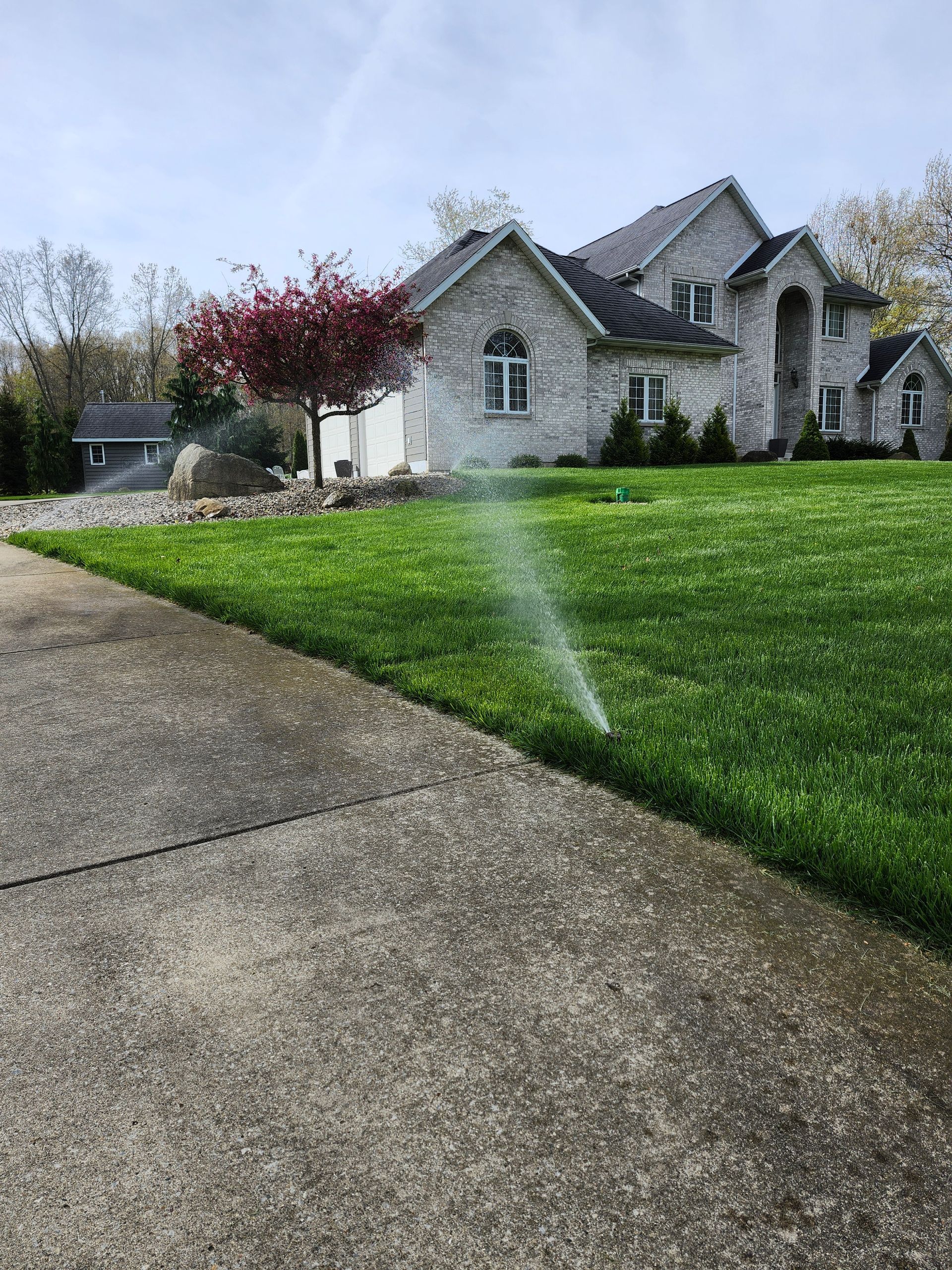 A lawn sprinkler is spraying water on a lush green lawn in front of a large house.
