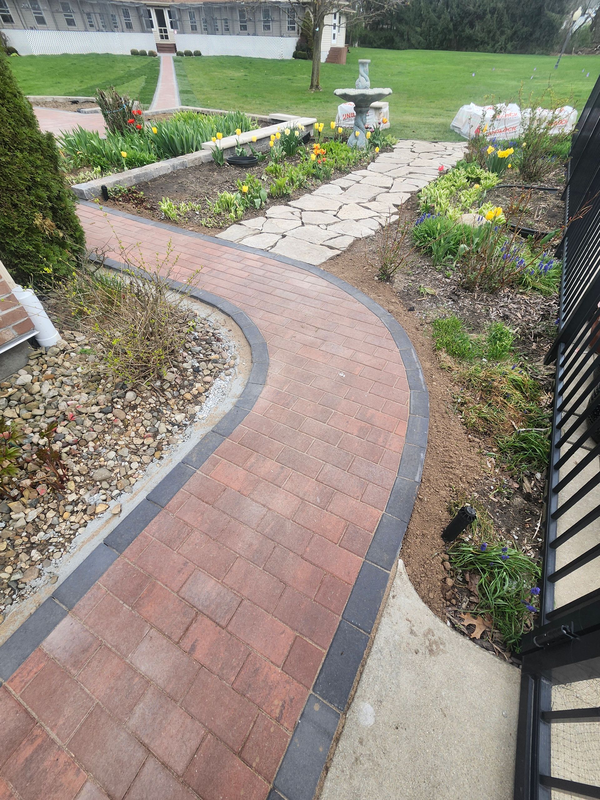 A brick walkway leading to a garden with a fountain in the middle.