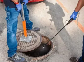 Workers in blue gloves use tools to inspect a manhole on a street.
