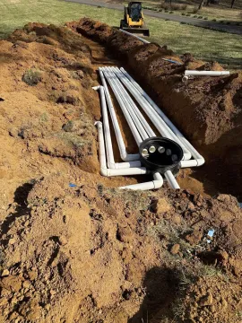 Trenched ground with white PVC pipes and a black junction box laid in an excavated ditch.
