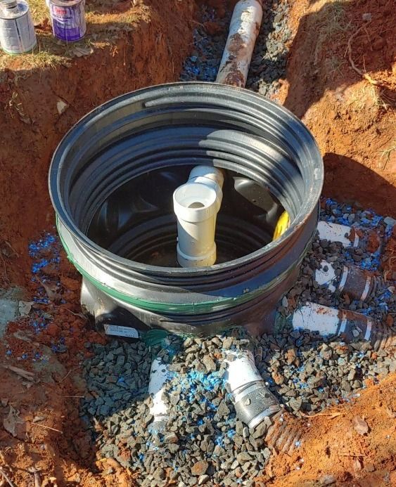 Large black corrugated pipe fitting with a white PVC pipe inside, set in a muddy excavation site.