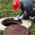 Worker in red hard hat inspecting an open manhole beside a grassy roadside.