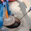Worker lifting a manhole cover beside an open sewer access hole on a sidewalk