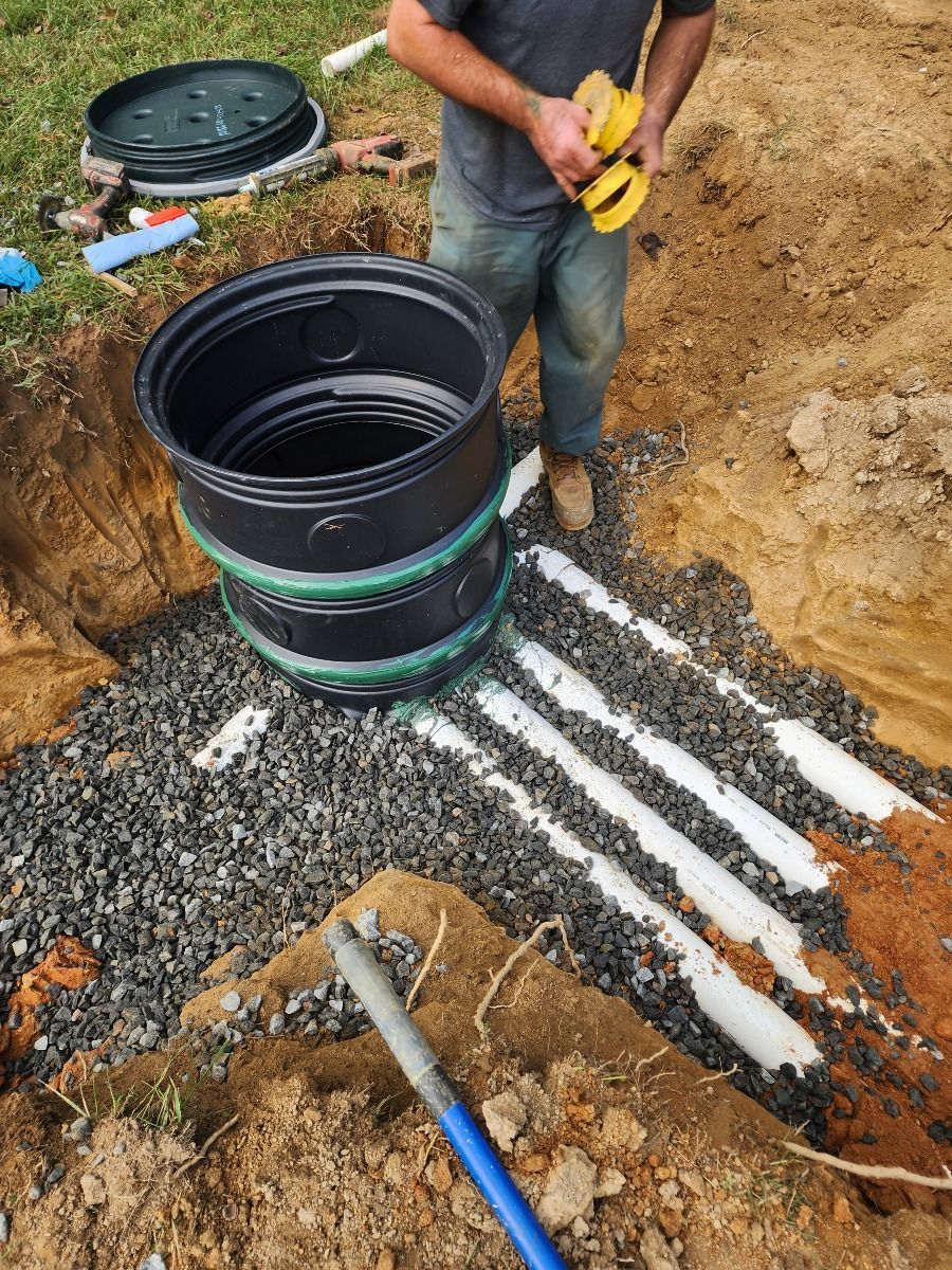 Worker installing black drainage pipe in a dirt trench with white pipes nearby.