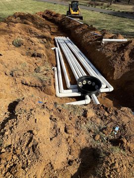 Trenched ground with white PVC pipes and a black junction box laid in an excavated ditch.