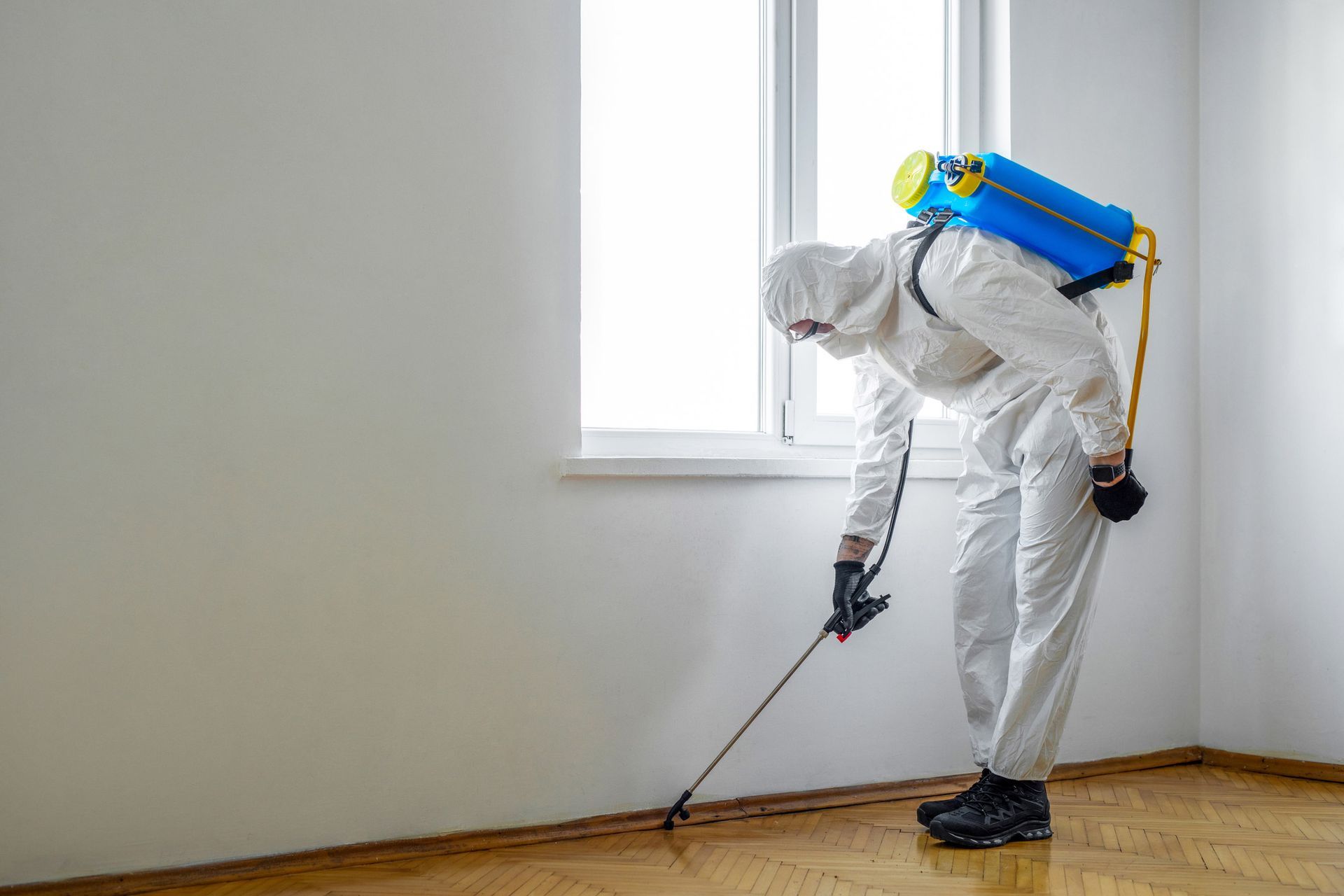 Pest technician wearing protective suit applying treatment to wall edges for pest control services.