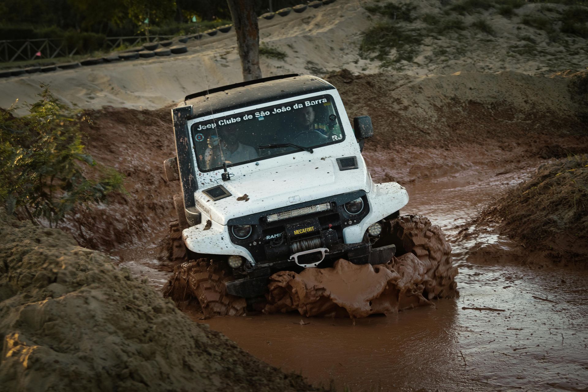 A white jeep is driving through a muddy area.