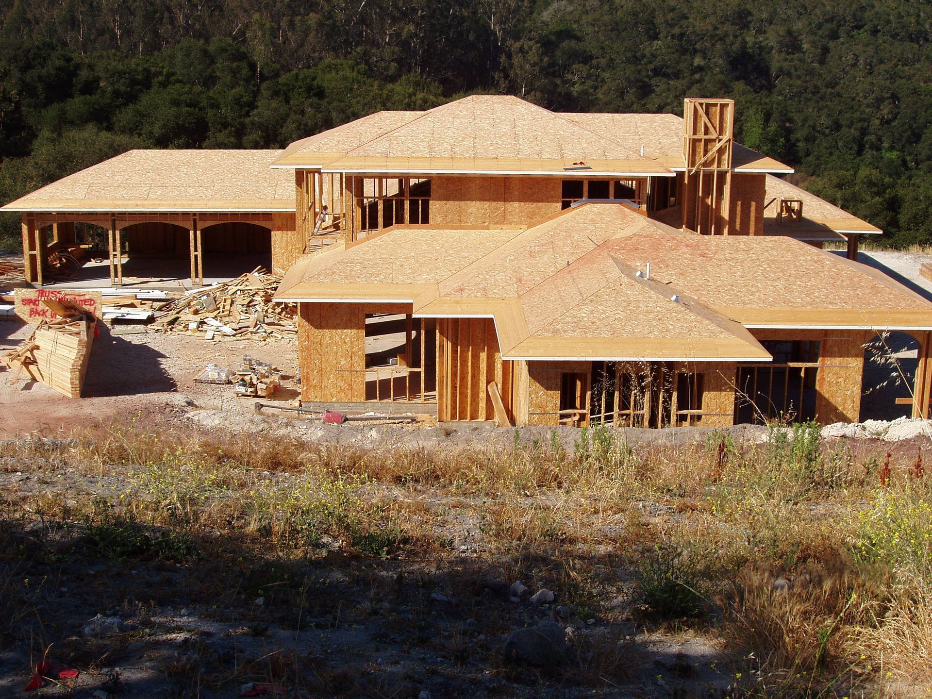 A house under construction with wooden framework, brown roof, set outdoors with trees in the background.