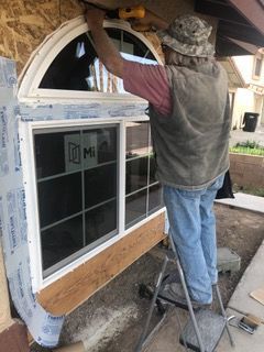 Man on ladder installing a window, wearing a hat and vest, outside a house.
