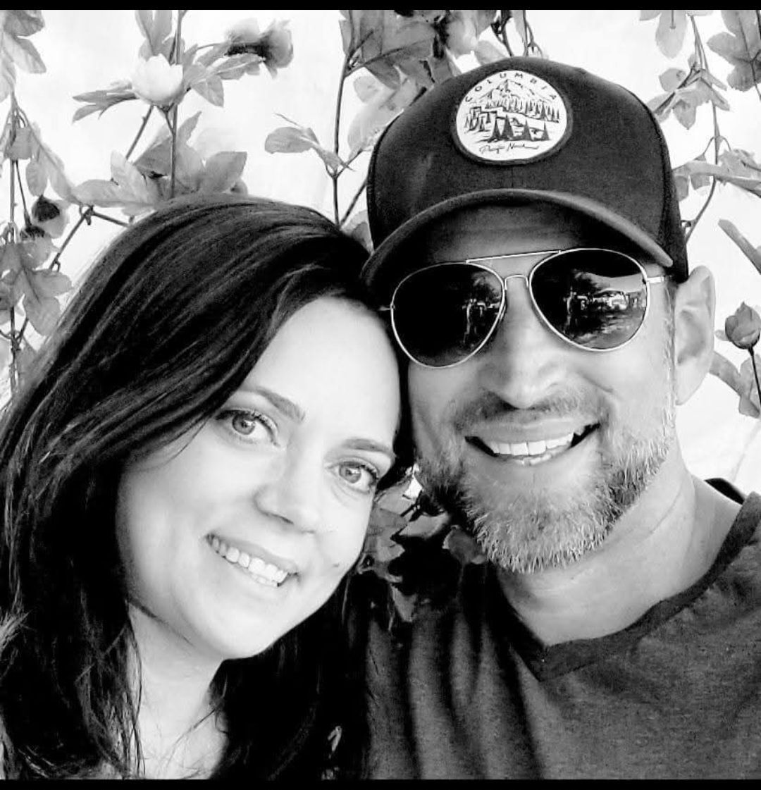 Smiling couple, black and white photo. Man wears a hat and sunglasses, woman has dark hair. Outdoor setting.