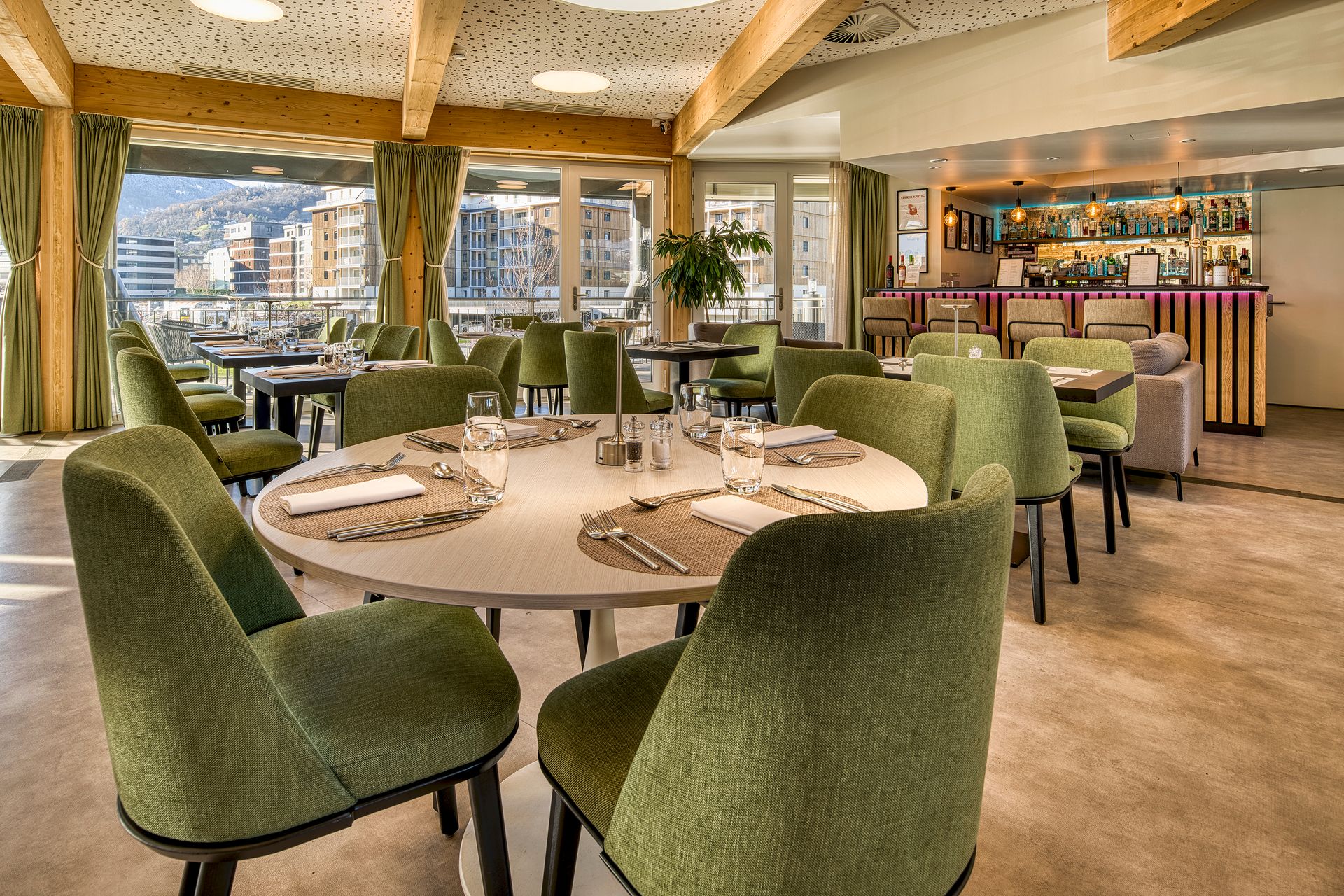 Interior view of a restaurant with round tables, green chairs, and a bar in the background. Windows offer city view.