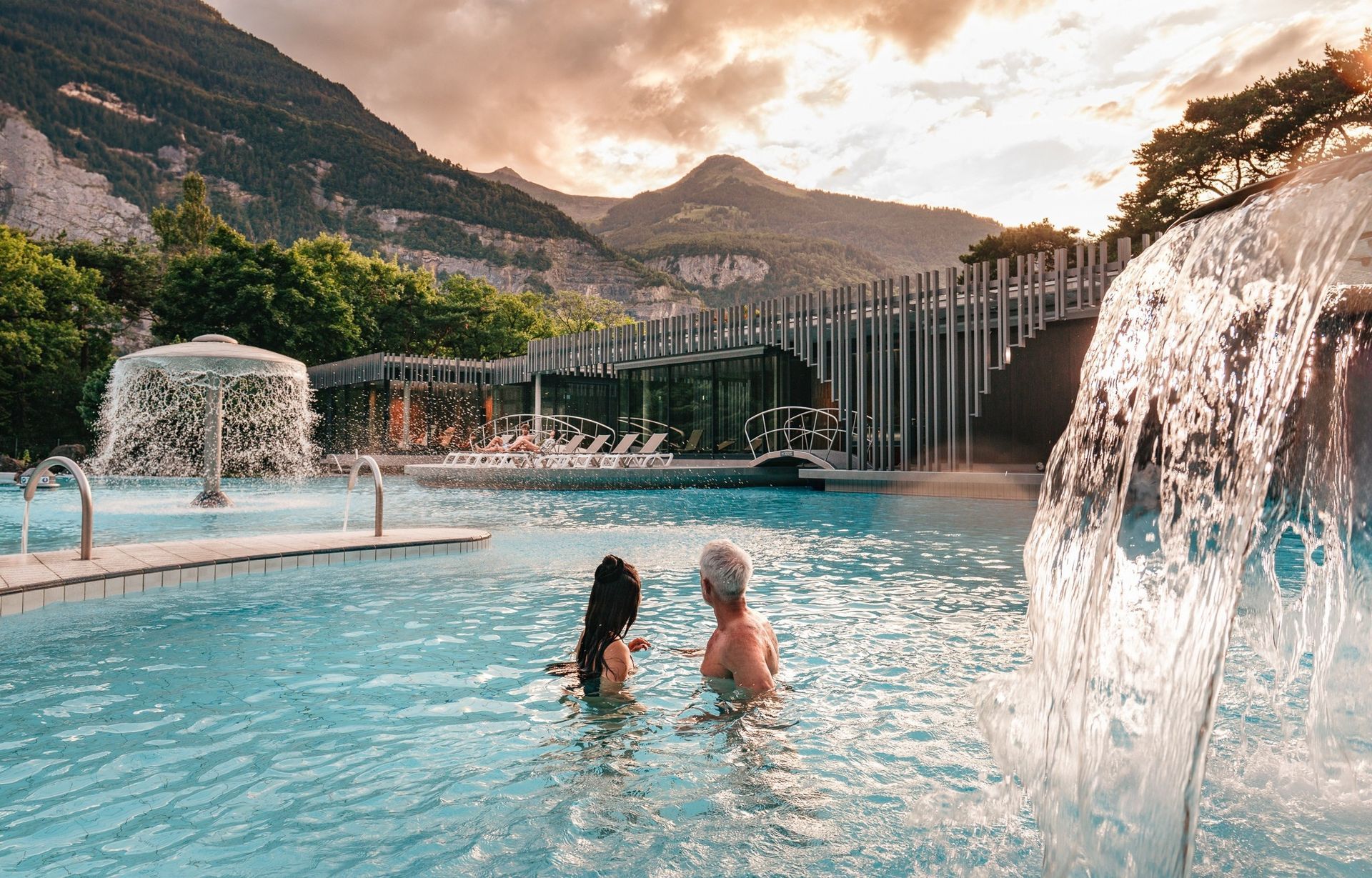 Two people in a turquoise pool with waterfall features and mountains at sunset