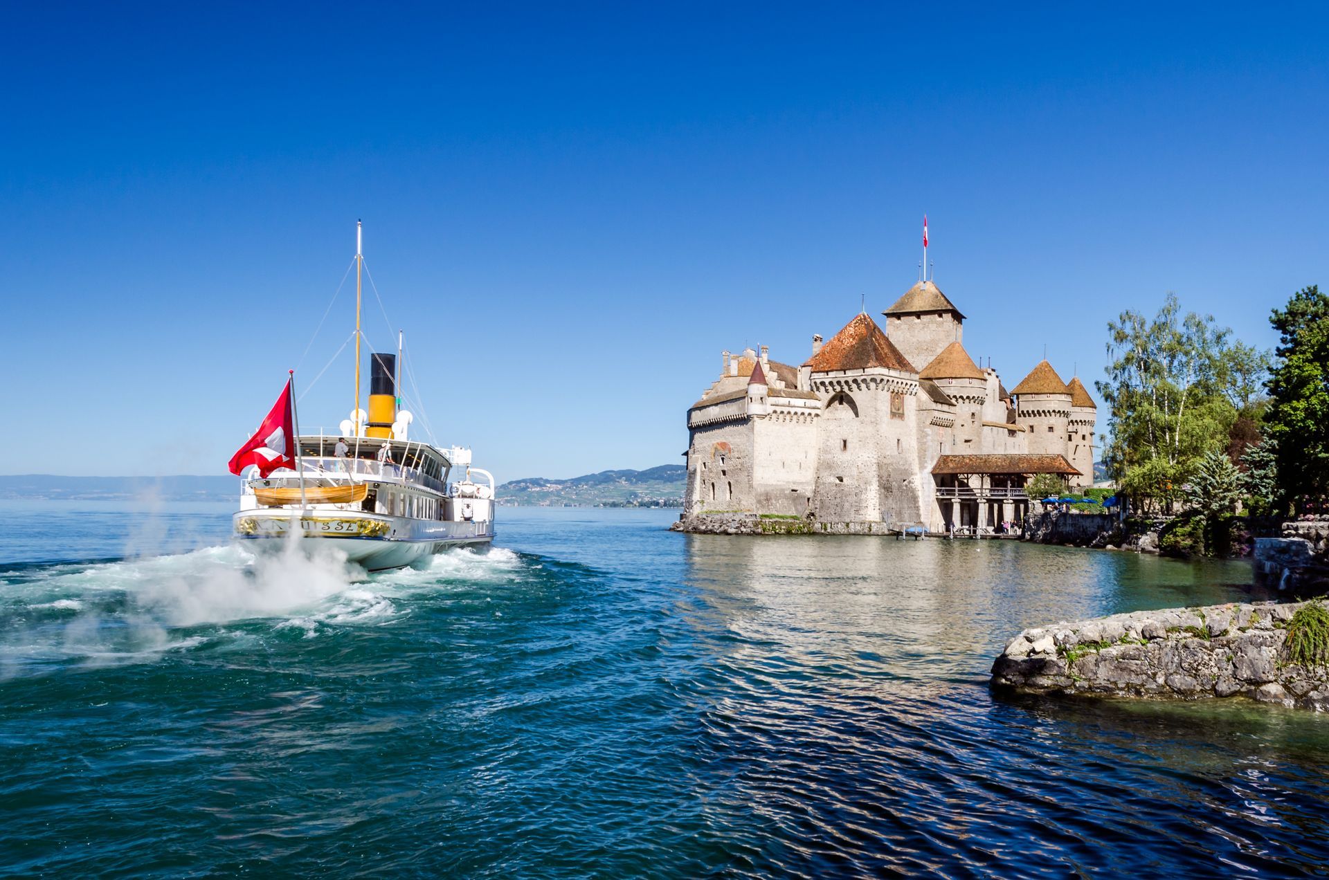 Red tour boat speeding past a stone castle on a blue lake under a clear sky