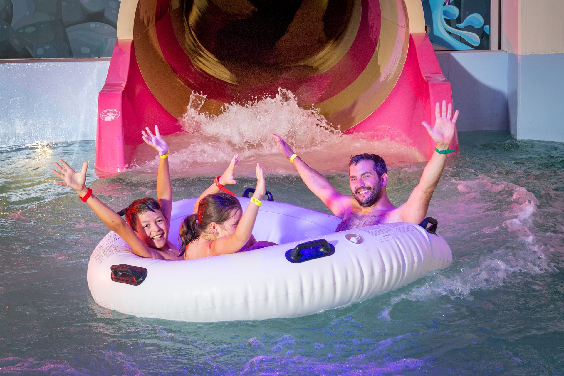 Une famille de trois personnes sur une bouée de toboggan aquatique, sortant dans la piscine. Toboggan rose et marron, mains levées, visages excités.