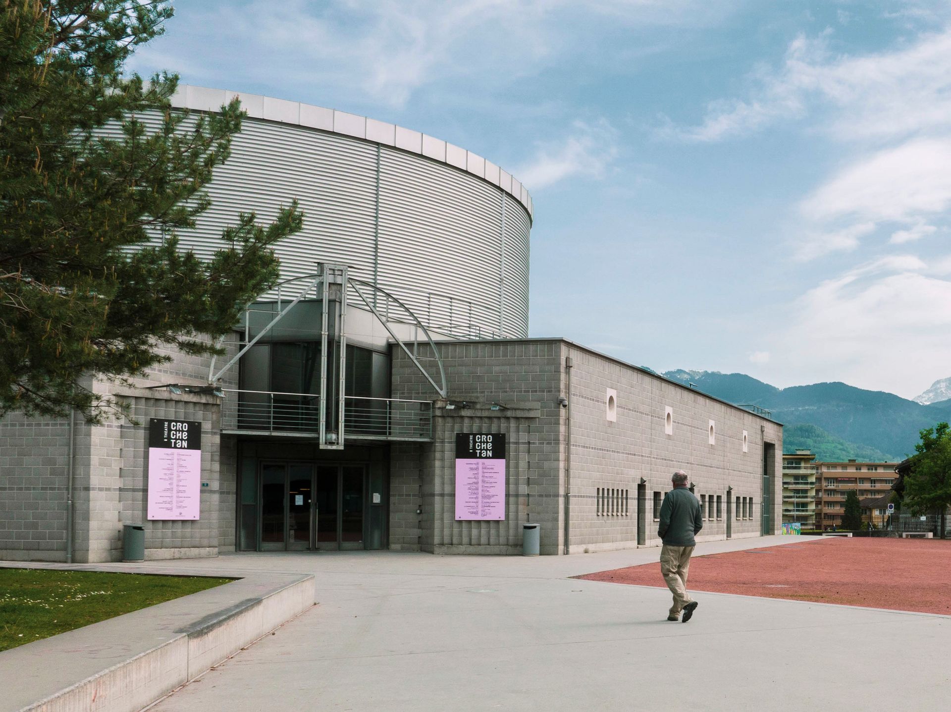 Modern building entrance with curved striped roof, pink banners, and a person walking on a paved plaza.