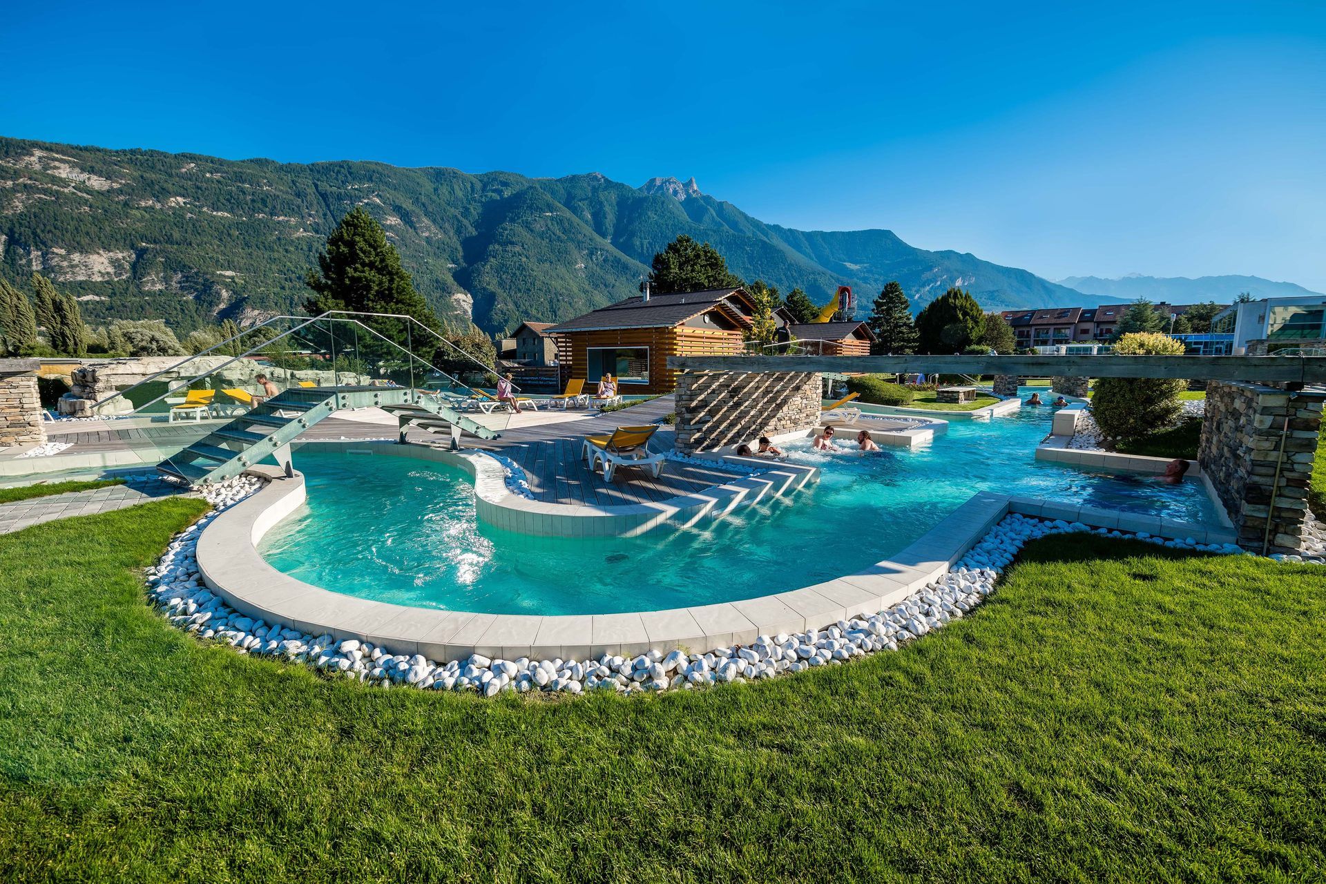 Luxury resort pool with curved turquoise water, lounge chairs, green lawn, and mountain backdrop