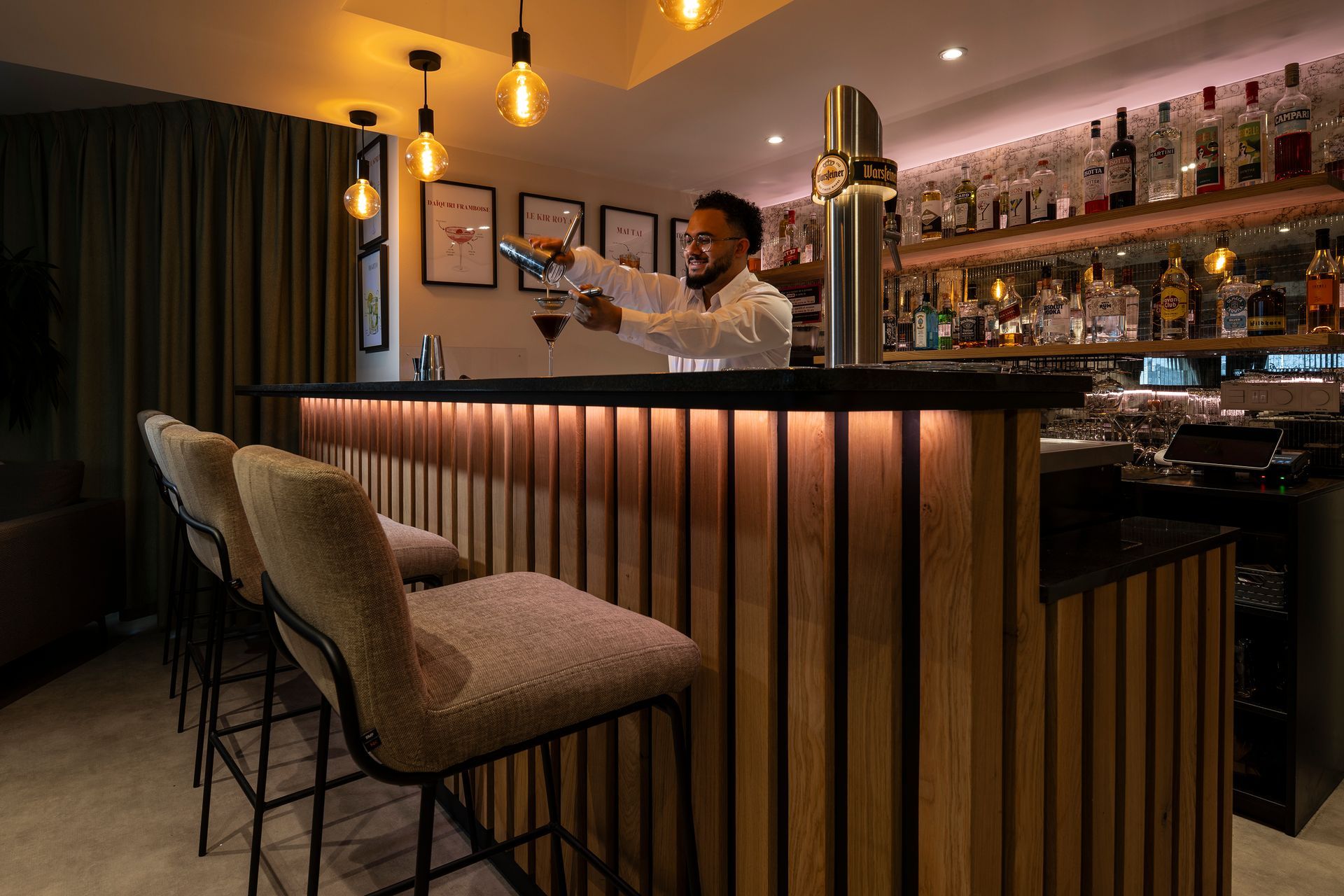 Ein Barkeeper schenkt an einer Bar mit Barhockern einen Drink ein. Die Barfront aus Holz ist warm beleuchtet.