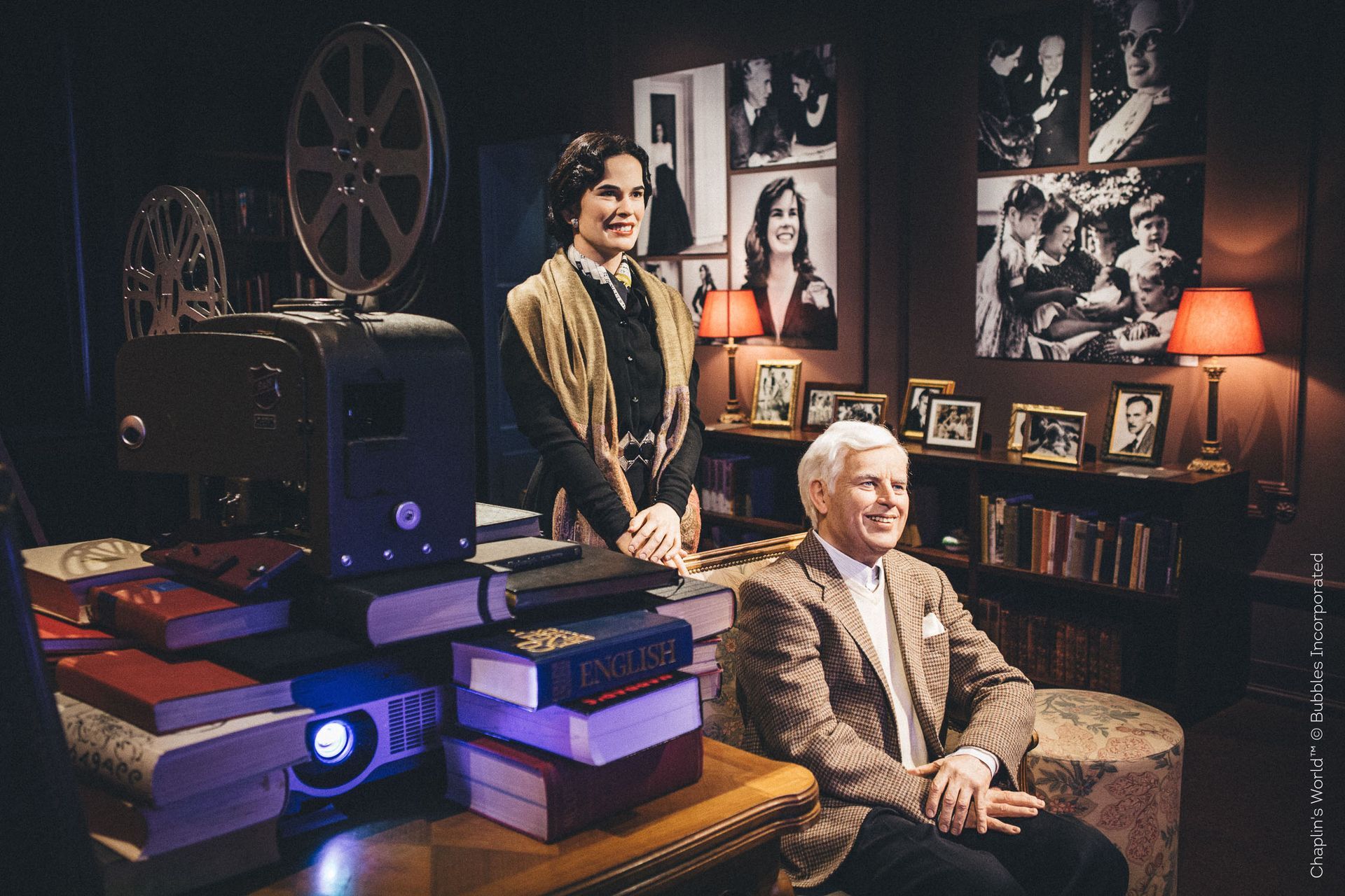 Two people in a cozy, retro room with books, lamps, and a film projector, one seated and one standing nearby