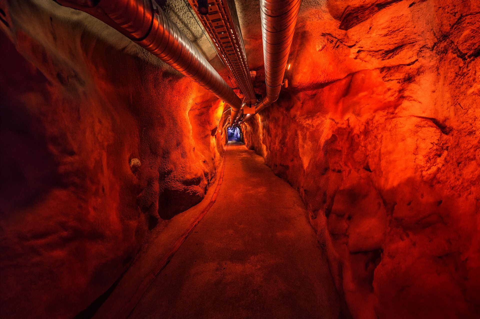 Narrow tunnel lit in red-orange with rocky walls and a walkway leading into the distance