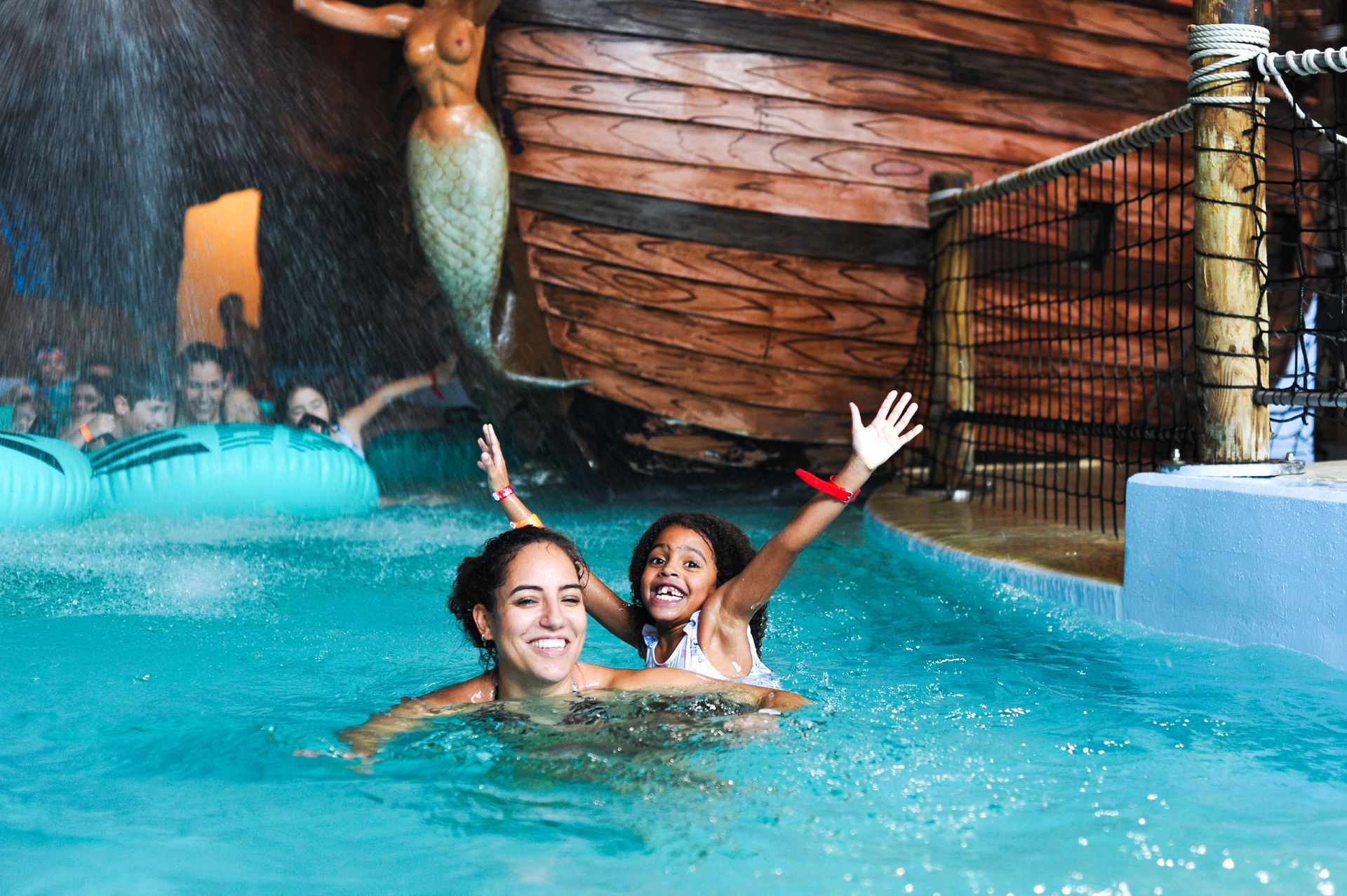 Two people swimming in a turquoise indoor pool near a mermaid statue and wooden boat decor