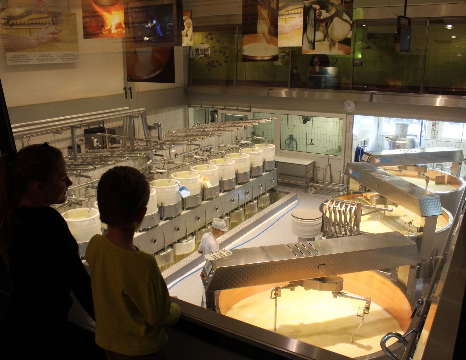 Two people overlook a miniature factory display with conveyor belts and machinery in a museum exhibit.