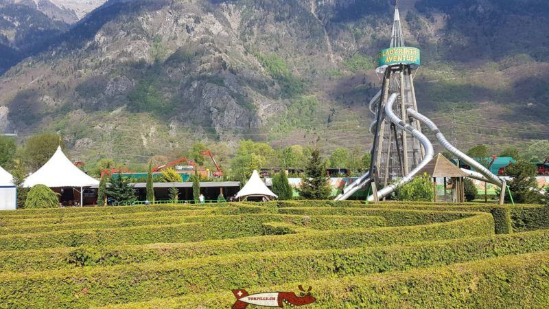 Theme park with a large spiral waterslide, white tents, and mountains in the background.
