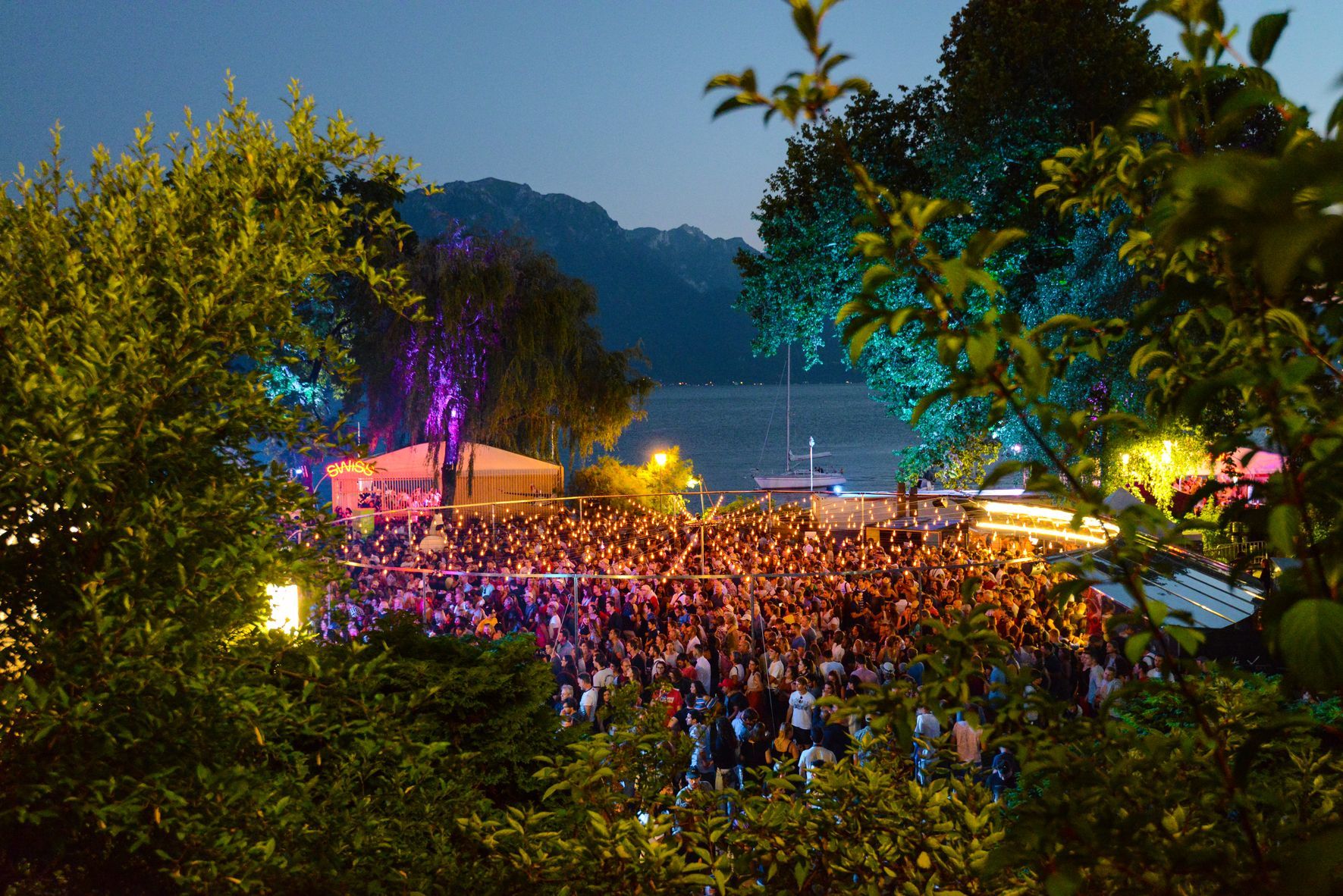 Große Menschenmenge bei nächtlicher Open-Air-Veranstaltung mit Bühne und Beleuchtung in der Nähe von Wasser und Bäumen.