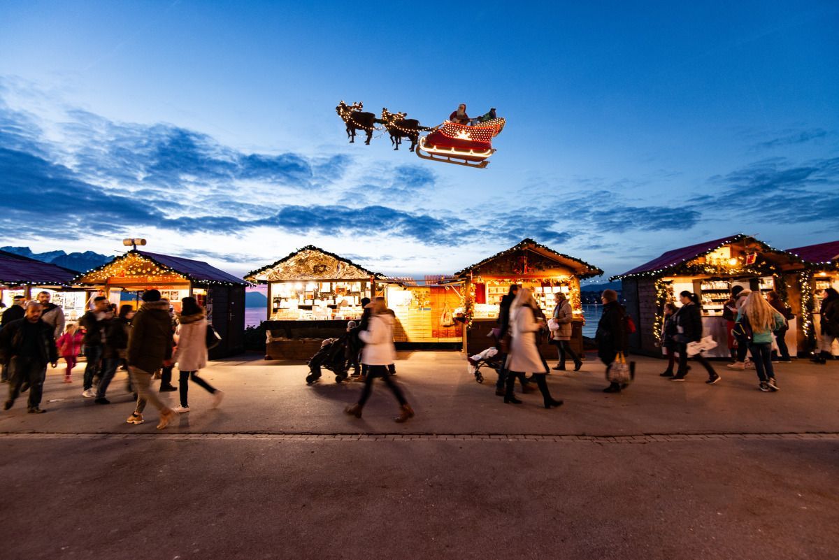 Der Schlitten des Weihnachtsmanns schwebt in der Abenddämmerung über einem festlich geschmückten Weihnachtsmarkt. Die Menschen kaufen an den beleuchteten Holzständen ein.