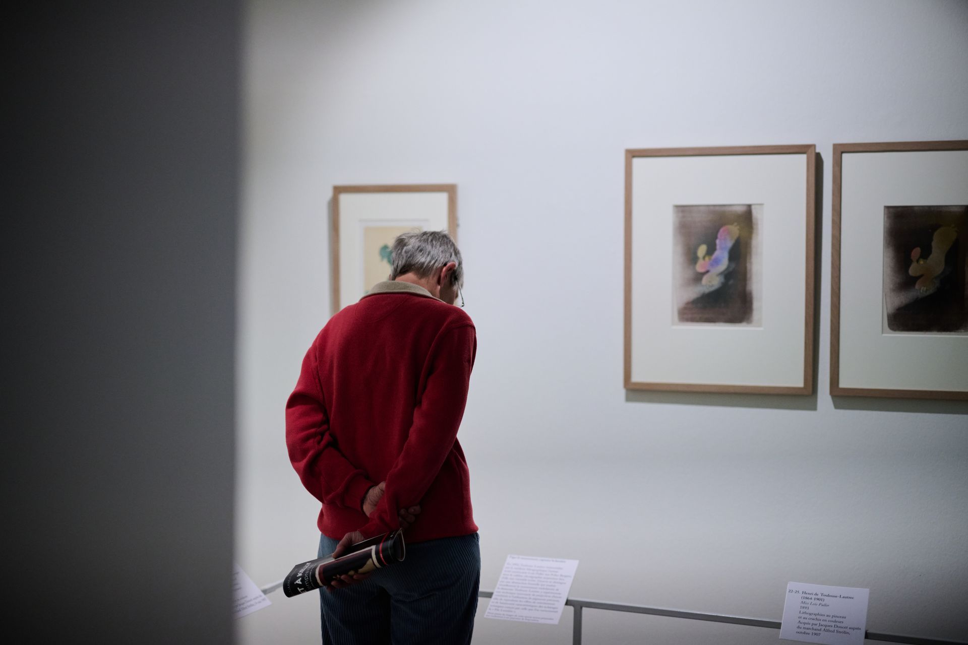 Person in a red sweater stands in a gallery, looking at framed artwork on a white wall.