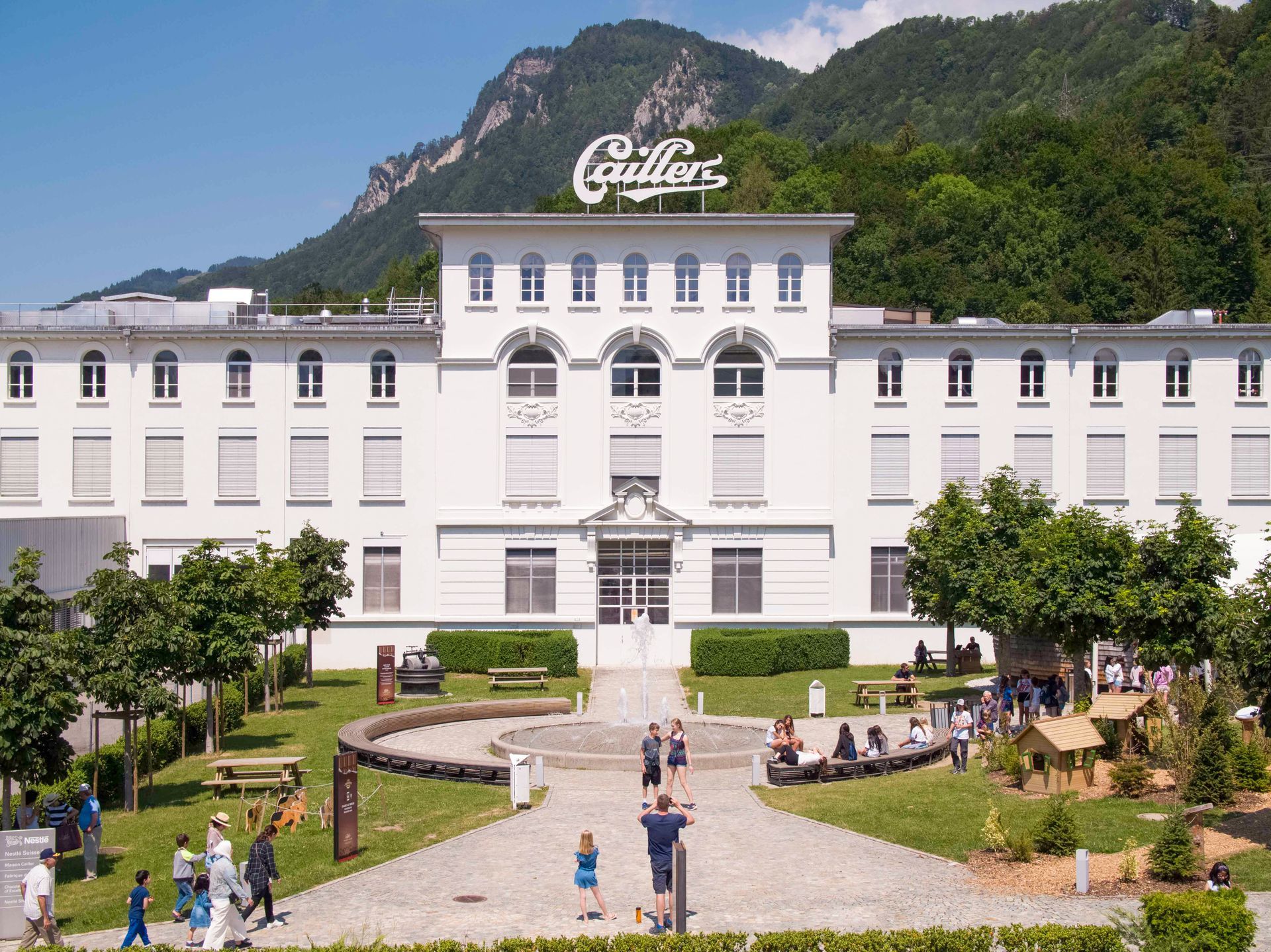 White hotel with central tower, green mountains behind, and people walking in the front garden.