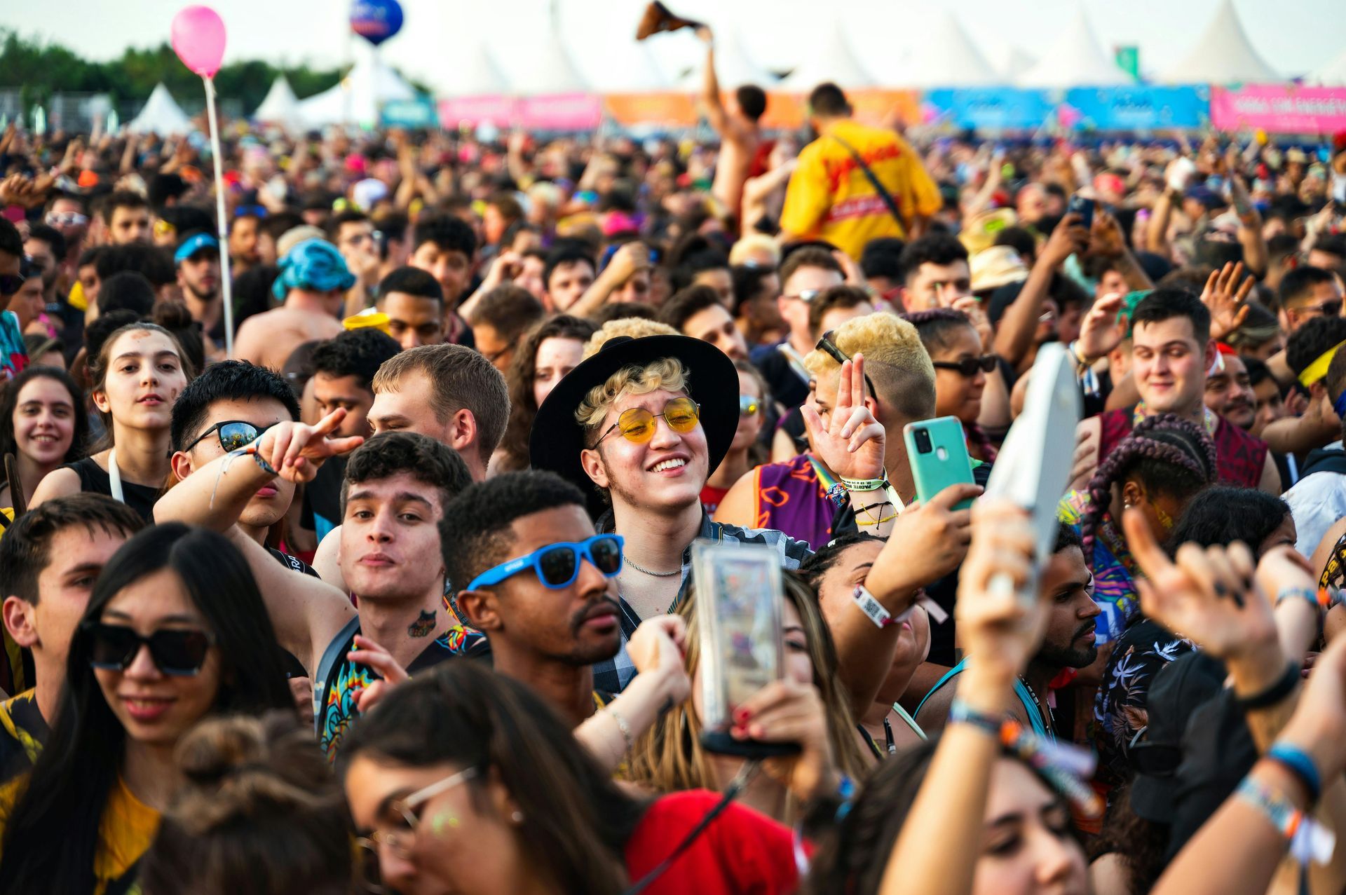 Große, bunte Menschenmenge bei einem Open-Air-Musikfestival; viele schauen nach oben, lächeln und machen Fotos.