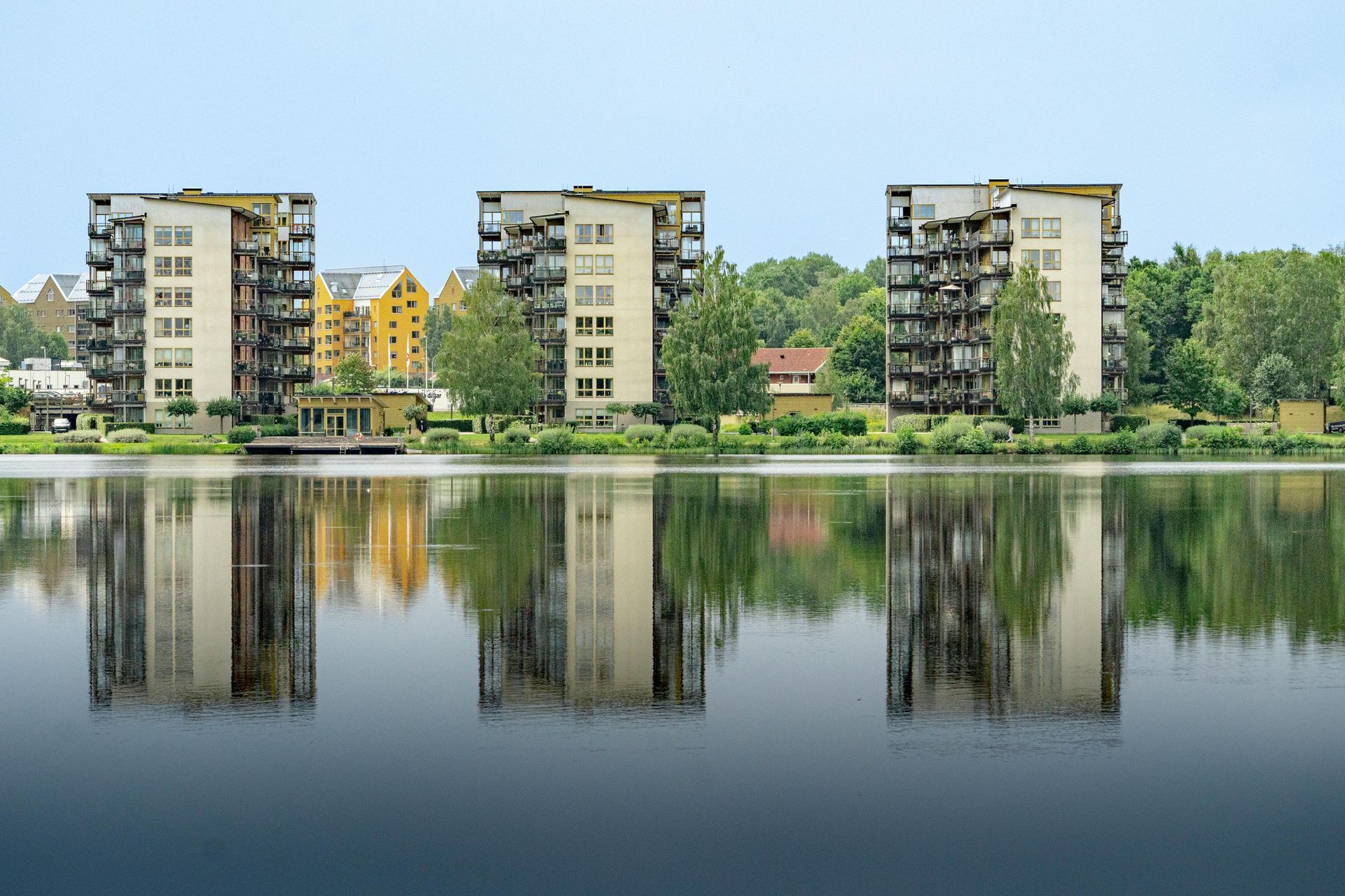 Wohnhäuser spiegeln sich im ruhigen See unter klarem Himmel, Bäume säumen das Ufer.