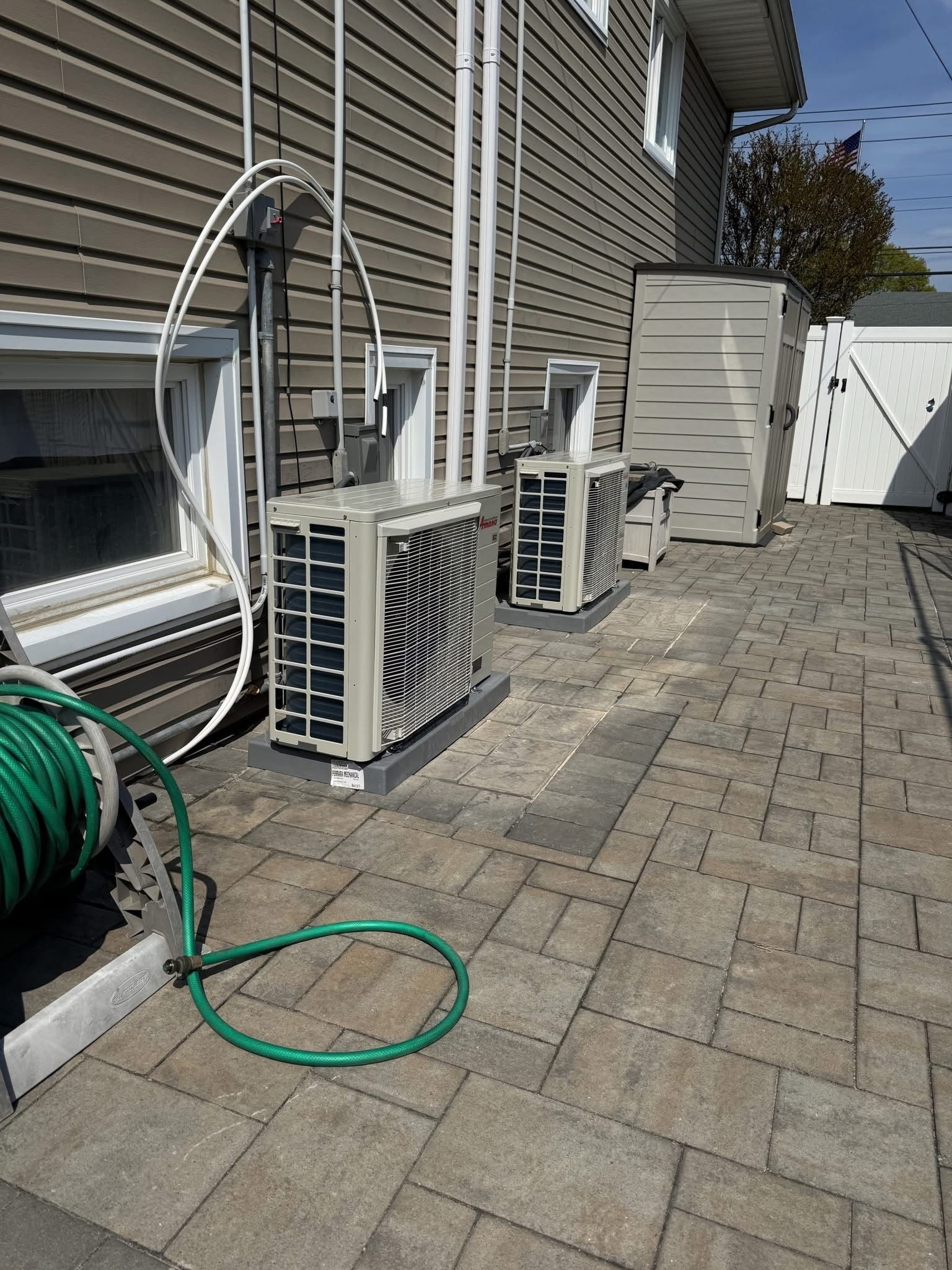 A gray central air conditioning unit sits on a concrete pad against a red brick exterior wall with decorative landscaping.