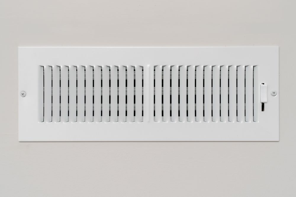 A white rectangular wall air vent with a manual slider control mounted on a plain wall.