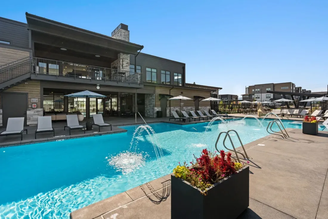 Outdoor apartment pool with lounge chairs, fountain jets, and a modern building under a clear blue sky.