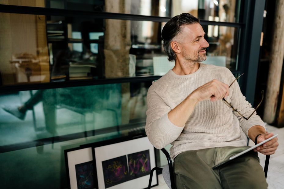 A person in a beige sweater holds a tablet and glasses, sitting in a studio-like space with framed art in the foreground.