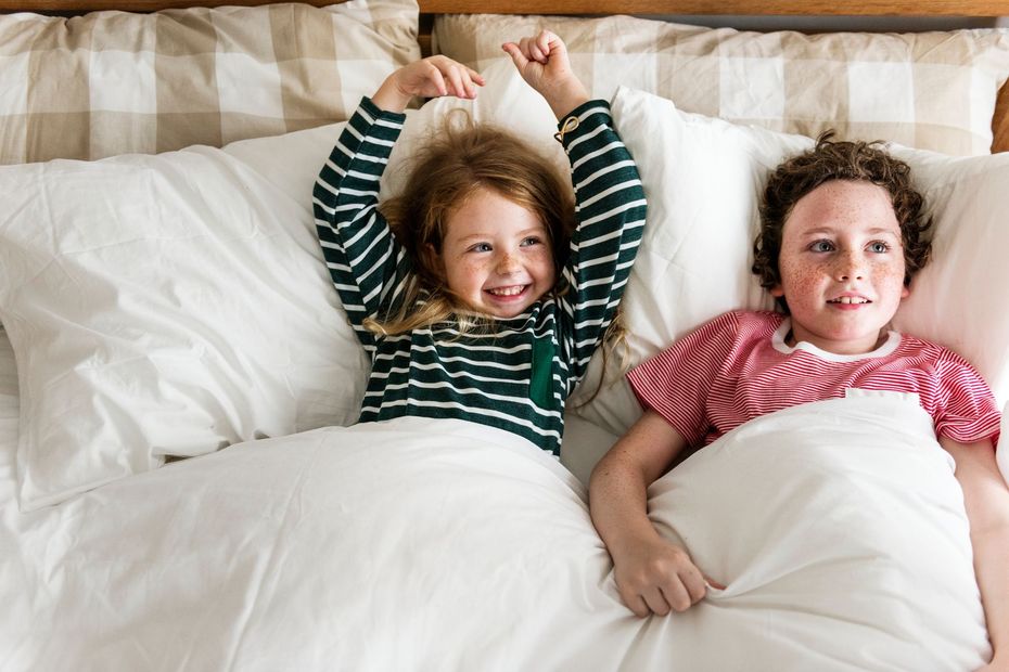 Two children sitting up in a white bed; one wears a green striped shirt with arms raised, the other wears a red shirt.