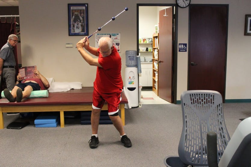 Person in red swings a golf club in a rehab room while others sit on a bench nearby.