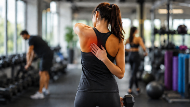 Woman stretching shoulder in a gym, with exercise equipment and a blurred person in the background