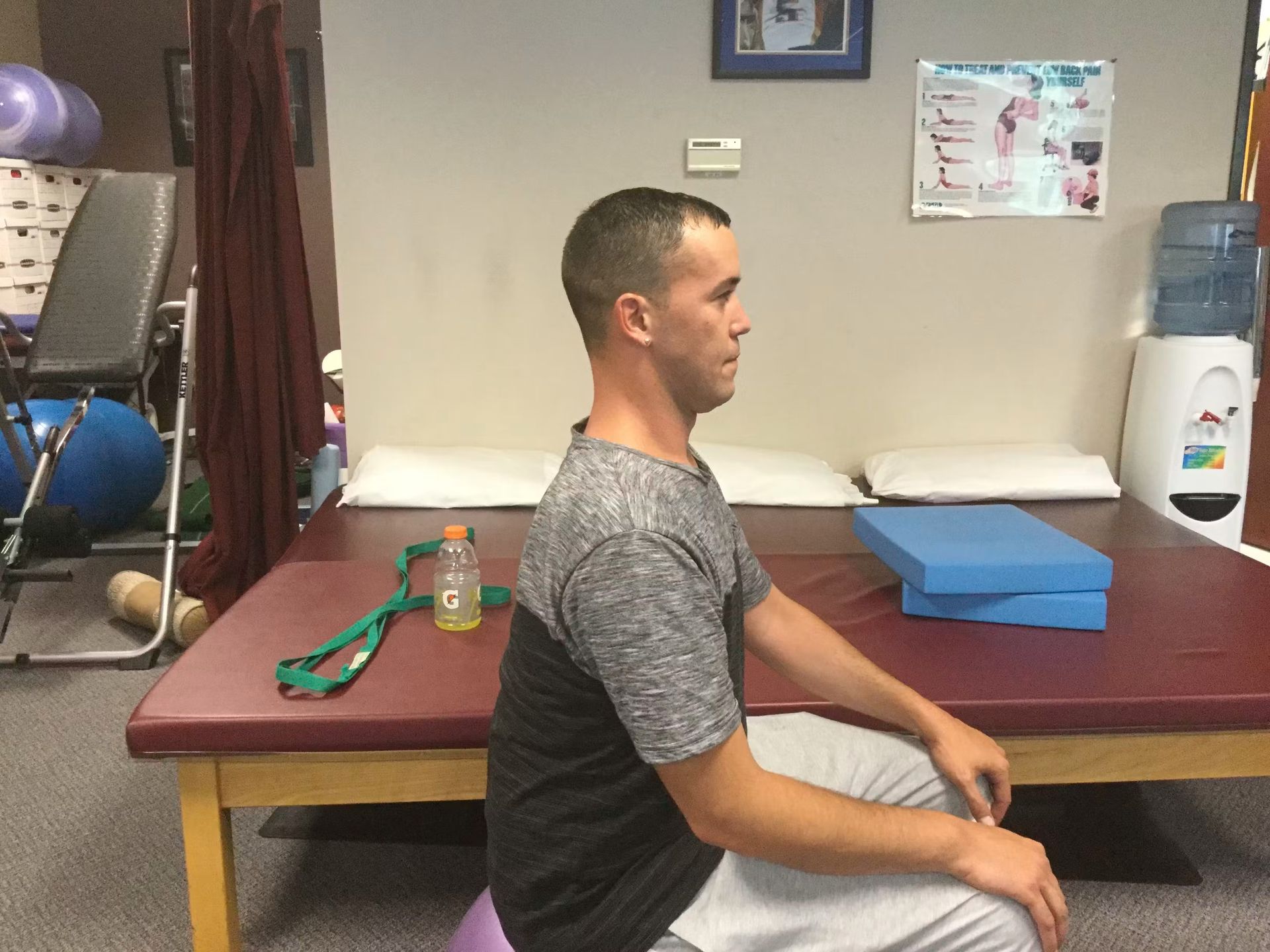 Man sitting upright on an exam table in a clinic room, facing right, with a resistance band nearby.