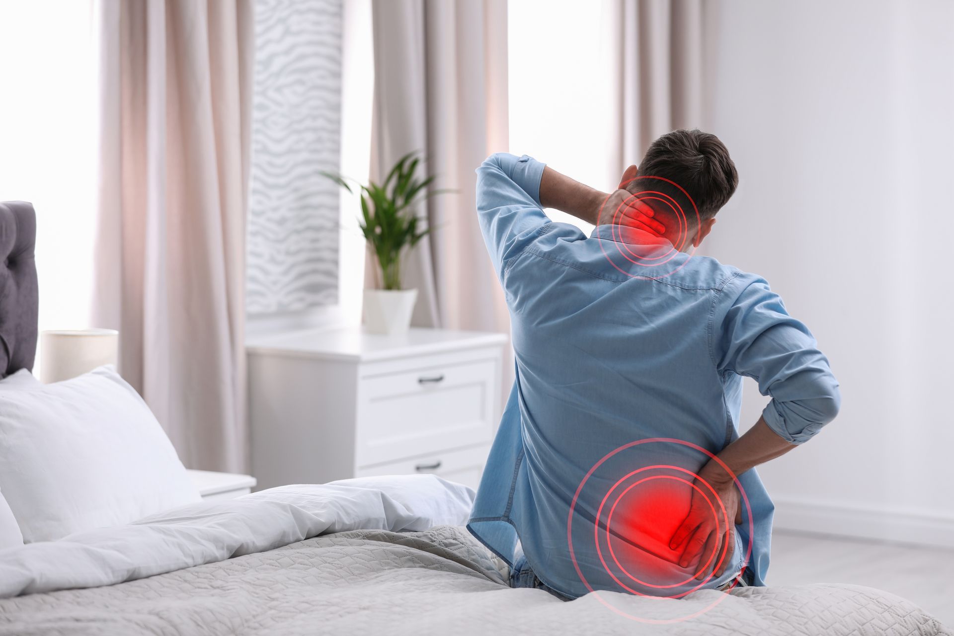 Person sitting on bed holding neck and lower back, with red pain highlights in a bedroom