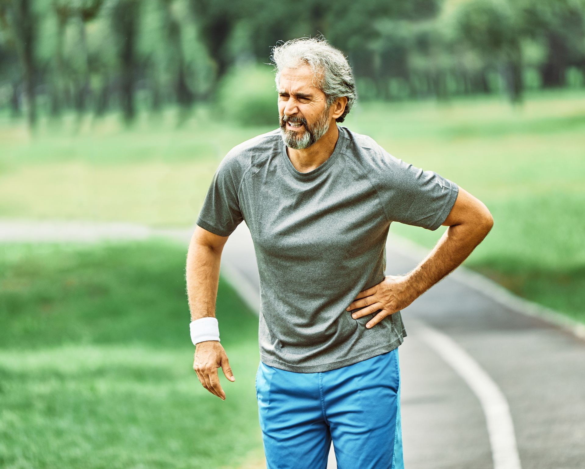 Man in gray T-shirt and blue shorts pausing on a park path, hand on hip, looking tired
