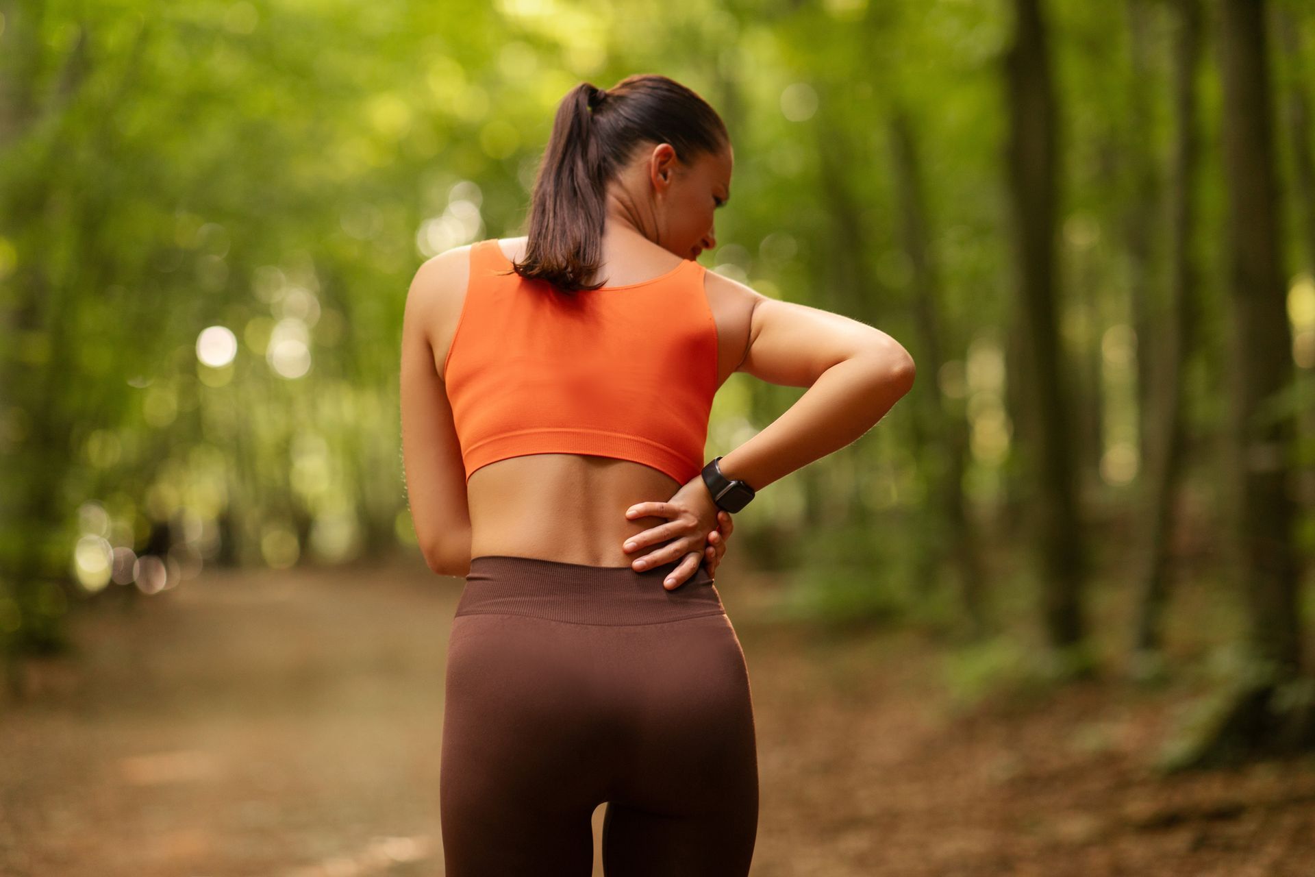 Woman in orange top and brown leggings stretching on a wooded trail, seen from behind.